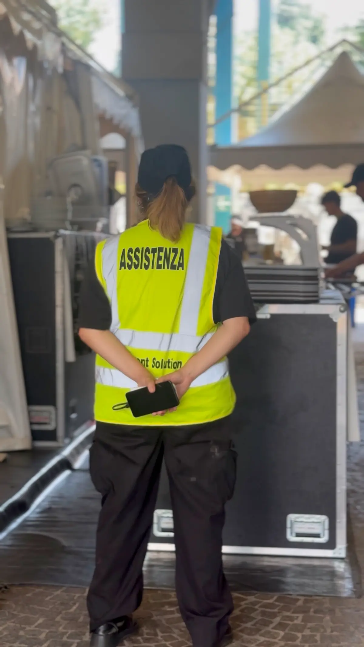 Un'operatrice YES con la pettorina Assistenza, di spalle, attende nel backstage tra i flight case